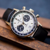 A Hamilton American Classic Intra-Matic Auto Chrono 40 mm Watch H38416711 by Hamilton Watches with a white dial, three black subdials, date at 6 o’clock, black tachymeter bezel, and black leather strap rests on a wooden surface.