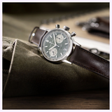 A close-up of the Hamilton Watches American Classic Intra-Matic Auto Chrono 40mm H38416560, featuring a green dial, silver case, and brown leather strap, resting on olive green fabric with a blurred notebook in the background.
