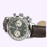 A Hamilton Watches American Classic Intra-Matic Auto Chrono 40mm H38416560 with a green panda dial, cream subdials, stainless steel case, and brown leather strap is displayed at an angle on a light purple background.