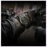 A close-up of a person wearing dark gloves and the Hamilton Watches Call of Duty: Modern Warfare 4 Khaki Field H69469930 Special Edition wristwatch by Hamilton Watches, with a blurred urban street background and posters visible on a building wall.