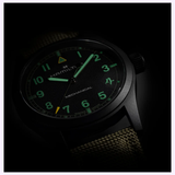 A Hamilton Watches Call of Duty: Modern Warfare 4 KHAKI FIELD H69469930 SPECIAL EDITION wristwatch by Hamilton Watches is displayed at an angle, featuring a black case, green luminescent numerals and hands, and a partially visible olive fabric strap in low light—evoking the rugged style of timepieces seen in Modern Warfare 4.