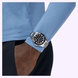 A person wearing a blue long-sleeve shirt and black pants has their left hand in their pocket, showcasing the Hamilton Watches H82415130 Khaki Navy Scuba Auto 40mm by Hamilton Watches with its sleek black dial.