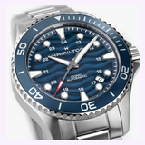 A close-up of the Hamilton Watches H82425140 Khaki Navy Scuba Auto 40 mm showcases its blue wave-patterned dial, silver hour markers, blue bezel, and stainless steel bracelet. The luminescent hands and markers exemplify Hamilton Watches’ renowned reloj de buceo craftsmanship.