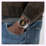 A person wearing the Hamilton Watches Jazzmaster Open Heart Auto 40 mm H32675181 by Hamilton Watches—with its refined automatic movement, silver band, and dark dial—has one hand in the pocket of blue jeans and is paired with a brown jacket featuring a knit cuff.
