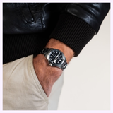 A person in a black leather jacket and light pants has their left hand in their pocket, showing a Hamilton Watches Khaki Aviation Pilot Auto H76215130 with a black dial and metal bracelet on their wrist.