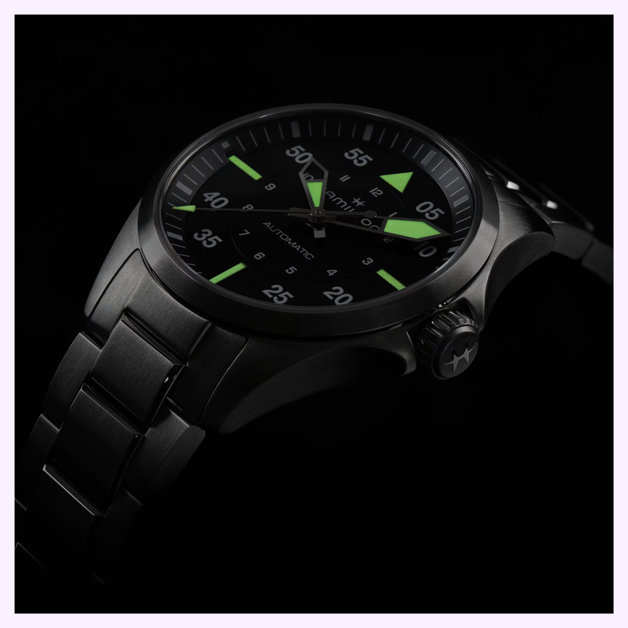 The Hamilton Watches Khaki Aviation Pilot Auto H76215130 features a black metal case and band, a round dial with luminous green hour markers and hands, all set against a dark backdrop.