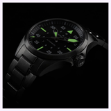 The Hamilton Watches Khaki Aviation Pilot Auto H76215130 features a black metal case and band, a round dial with luminous green hour markers and hands, all set against a dark backdrop.