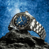 A Hamilton Watches Khaki Field Murph 38mm H70405140 in silver with a blue dial lies atop a dark rock, framed by a vivid, star-filled sky.