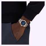 A person wearing a Hamilton Watches Khaki Field Murph 38mm H70405140 wristwatch by Hamilton Watches, featuring a silver case and blue face, has their hand in their pocket. They’re dressed in a dark long-sleeve top and blue pants, channeling the sleek style made famous by the Interstellar watch.