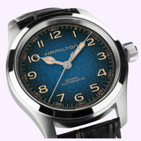 A close-up of the Hamilton Watches Khaki Field Murph 38mm H70405740 features a blue textured dial with gold numerals, a silver case, and a black leather strap. This sophisticated timepiece from Hamilton Watches is showcased displaying the time as 10:10:37.