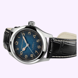A close-up of the Hamilton Watches Khaki Field Murph 38mm H70405740 wristwatch, featuring a black leather strap, black dial with white Arabic numerals, and a silver case, set against a light purple background. This striking automatic watch is crafted by Hamilton Watches.