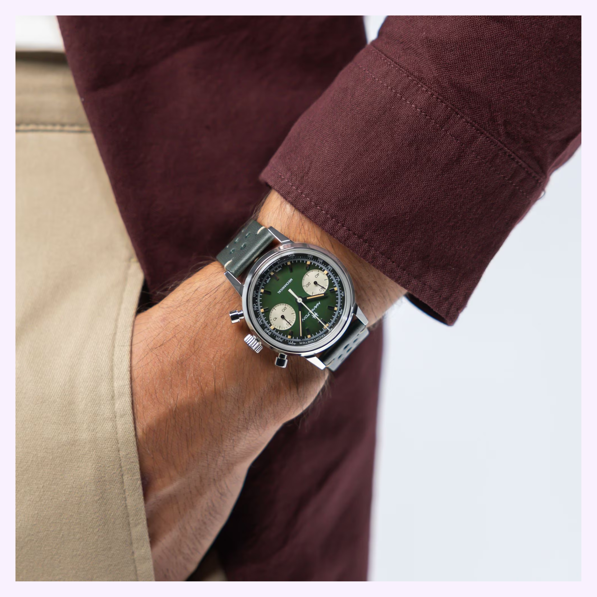 Wearing a maroon long-sleeve shirt and beige pants, a person has their hand in their pocket, displaying the Hamilton Watches American Classic Intra-matic Chronograph H38429861 with a green dial and cream subdials on a silver band.