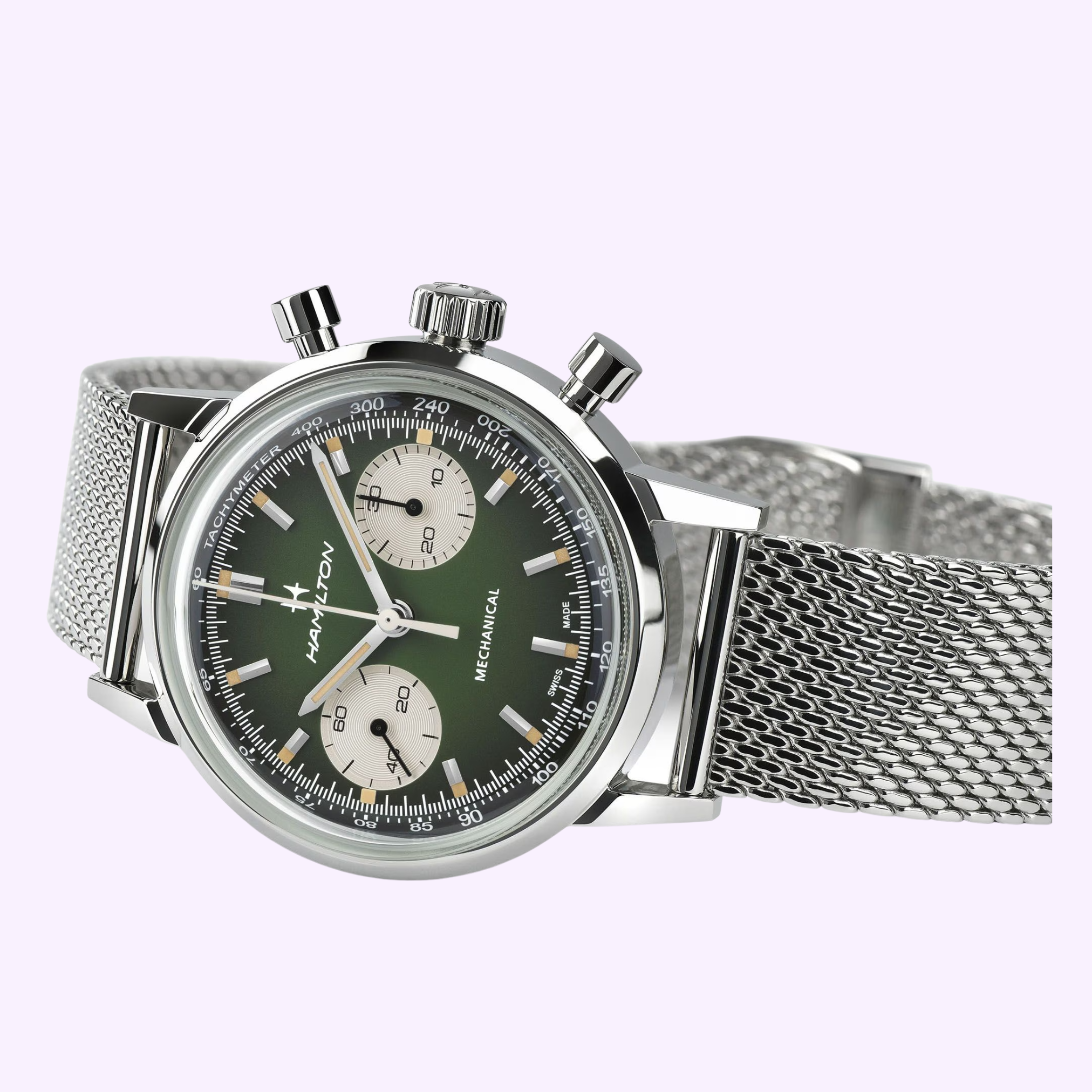 The Hamilton Watches American Classic Intra-matic Chronograph H38429861 features a silver case, green dial with two white subdials, silver hour markers, and a stainless steel mesh band, displayed on a light purple background.