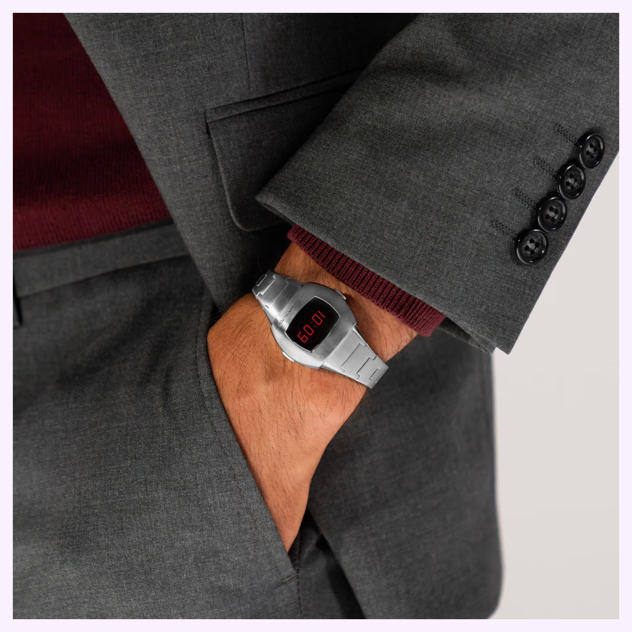 A person in a gray suit and maroon sweater, hand in pocket, showcases a Hamilton Watches American Classic PSR 74 H52304130 digital watch reading 06:01.
