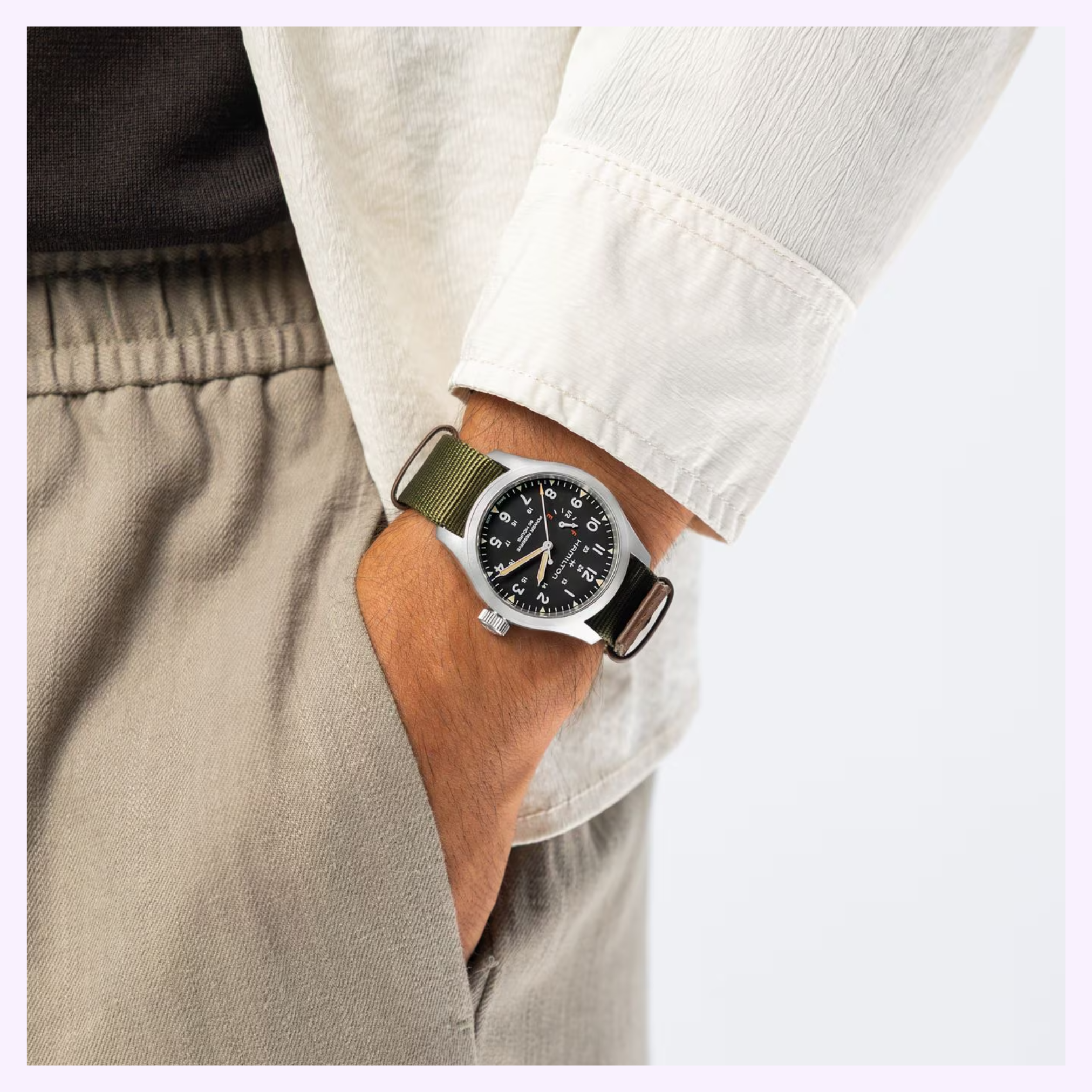 A person in beige pants and a white shirt, one hand in their pocket, showcases the Hamilton Watches Khaki Field Mechanical Power Reserve 40 mm H69509930 with an 80-hour reserve and green fabric strap on their left wrist.
