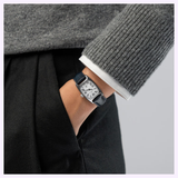 A person dressed in a grey sweater and black pants has their hand in their pocket, showcasing a women’s luxury timepiece—the Hamilton Watches American Classic Boulton Small Second Quartz H13321611, featuring a silver case, black leather strap, and a white dial with Roman numerals.