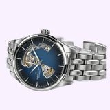 The Hamilton Watches Jazzmaster Open Heart Auto H32675140 is a silver automatic watch with a blue face, visible internal gears, and a metal link band, shown against a light purple background.