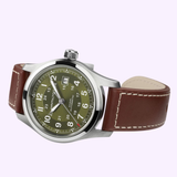 The Hamilton Watches Khaki Field Auto 42mm H70605560 is an automatic military watch with a brown leather strap, olive green dial, white numerals, silver case, date display, and both 12/24-hour markings.