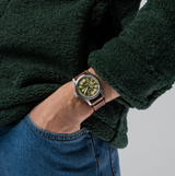 A person in a dark green fleece jacket and blue jeans, left hand in pocket, wears a Hamilton Watches Khaki Field Auto 42mm H70605560 with a green dial and brown leather strap.