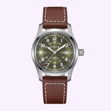 The Hamilton Watches Khaki Field Auto 42mm H70605560 features a stainless steel case, brown leather strap, olive green dial with white numerals, date window at 3 o’clock, and luminous hands, shown on a light purple background.