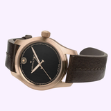 The Hamilton Watches Khaki Field Auto The Odyssey Limited Edition H70675530 features a bronze case, black dial with gold accents, Hamilton logo, hour markers, and a dark brown leather strap that contrasts with the light purple background.