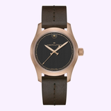 The Hamilton Watches Khaki Field Auto The Odyssey Limited Edition H70675530, featuring a bronze case, black textured dial with gold accents, and brown leather strap, is elegantly displayed on a light lavender background.