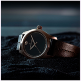 A close-up of the Hamilton Watches Khaki Field Auto The Odyssey Limited Edition H70675530, featuring a dark face, metallic case, and black leather strap, displayed on textured black fabric with a Christopher Nolan-inspired blurred backdrop.