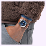 A person wearing a brown fleece jacket and gray pants has their hand in their pocket, revealing a Hamilton Watches Khaki Field King Day-Date 40mm H64475540 with a brown leather strap, blue dial, silver casing, and day-date display on their wrist.