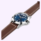 The Hamilton Watches Khaki Field King Day-Date 40mm H64475540, featuring a blue dial with white Arabic numerals and a silver case, is set against a pale background. Its brown leather strap with white stitching enhances the rugged design of this automatic field watch from Hamilton Watches, while the practical day-date display offers refined functionality.