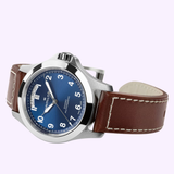 Introducing the Hamilton Watches Khaki Field King Day-Date 40mm H64475540, an automatic field watch featuring a striking blue dial, silver case, and a brown leather strap with white stitching. Crisp white numerals ensure easy readability, while its clear day-date display at the 12 o’clock position adds enhanced functionality.