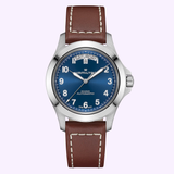 A Hamilton Watches Khaki Field King Day-Date 40mm automatic watch (model H64475540) in silver, featuring a blue dial with white numerals, day-date display, and a brown leather strap with white stitching, displayed against a light purple background.