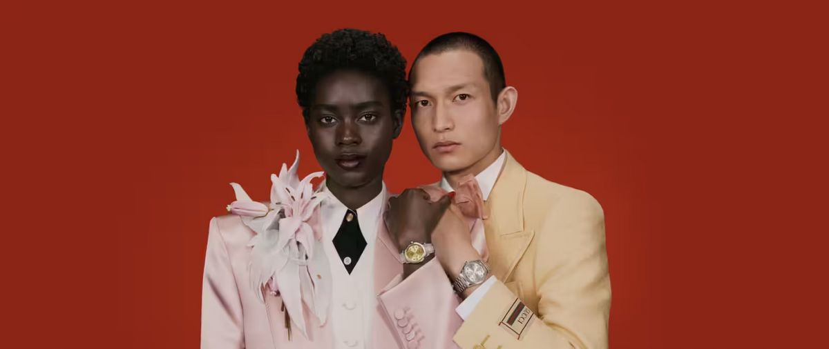 A woman in a light pink suit with floral details and a man in a pale yellow suit pose side by side against a solid red background. Both have serious expressions and are looking directly at the camera.