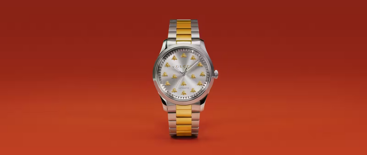 A silver and gold Gucci wristwatch with a metallic band, displayed upright against a solid orange background. The watch face features gold bee motifs in place of traditional numerals.