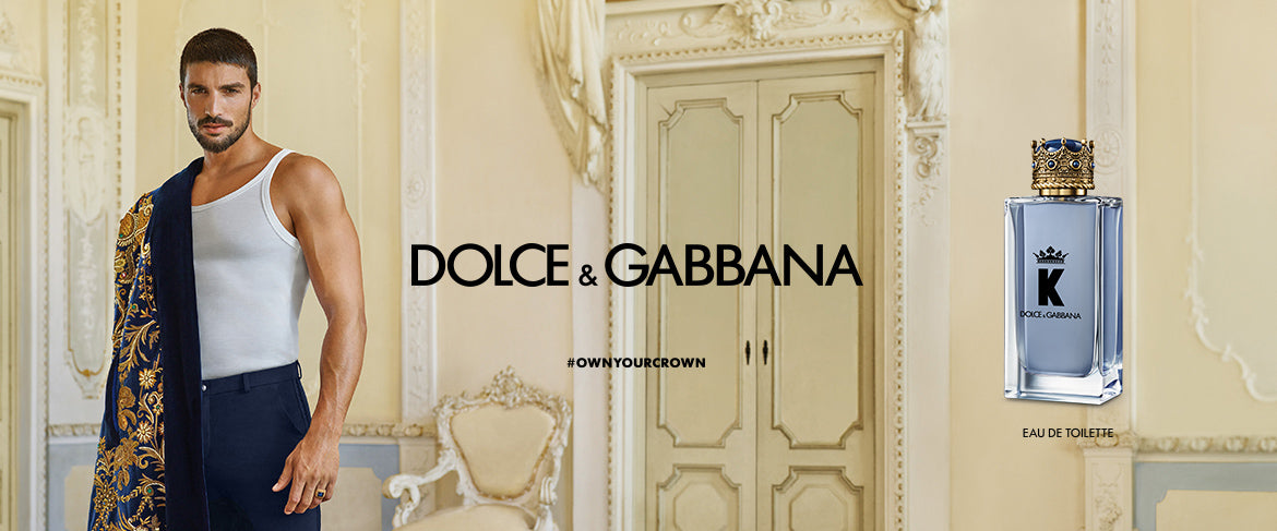 A man in a white tank top and dark pants stands in an ornate room, holding a blue and gold patterned jacket. Next to him is the Dolce & Gabbana logo and a bottle of K Eau de Toilette perfume.