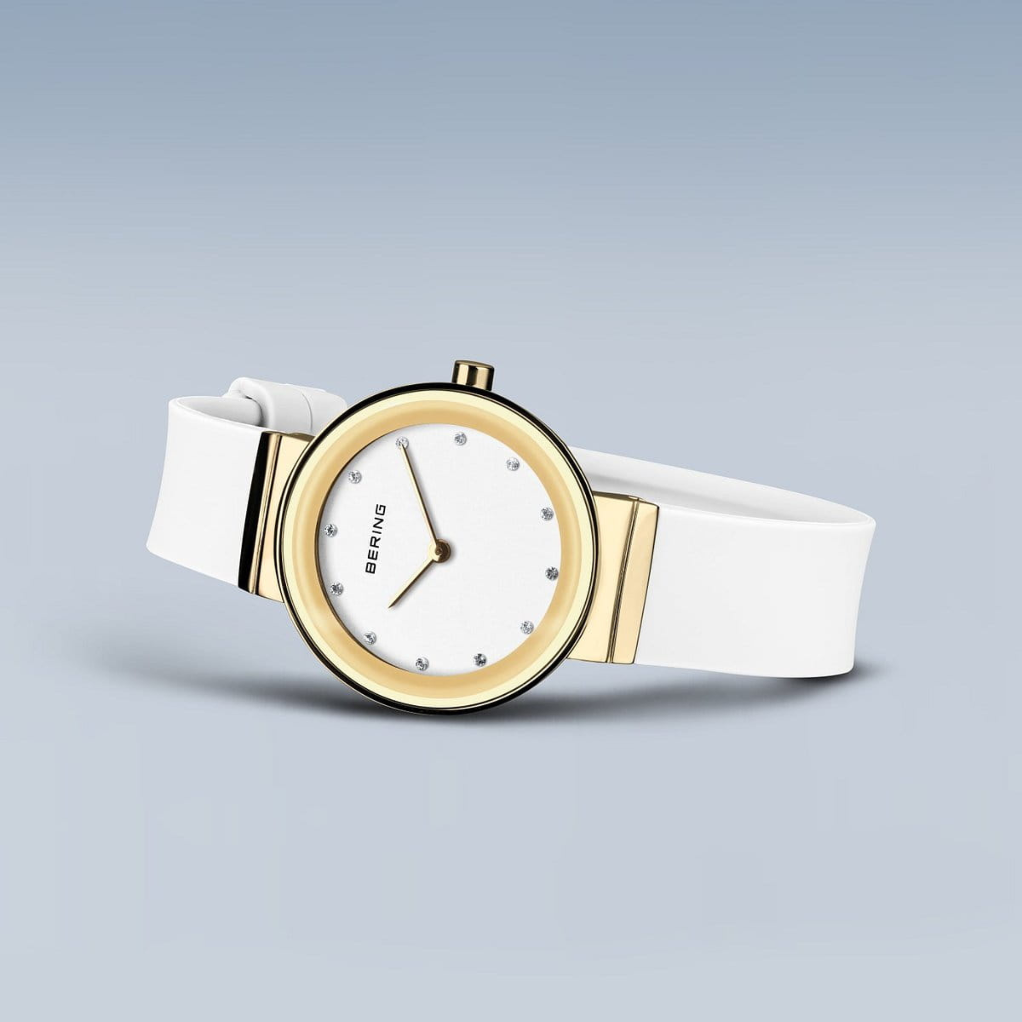 The Bering Watches Classic 10129-934 women's watch features a white band, gold-tone round stainless steel case, white dial with crystal hour markers, and the Bering Watches logo, shown on a light blue background.
