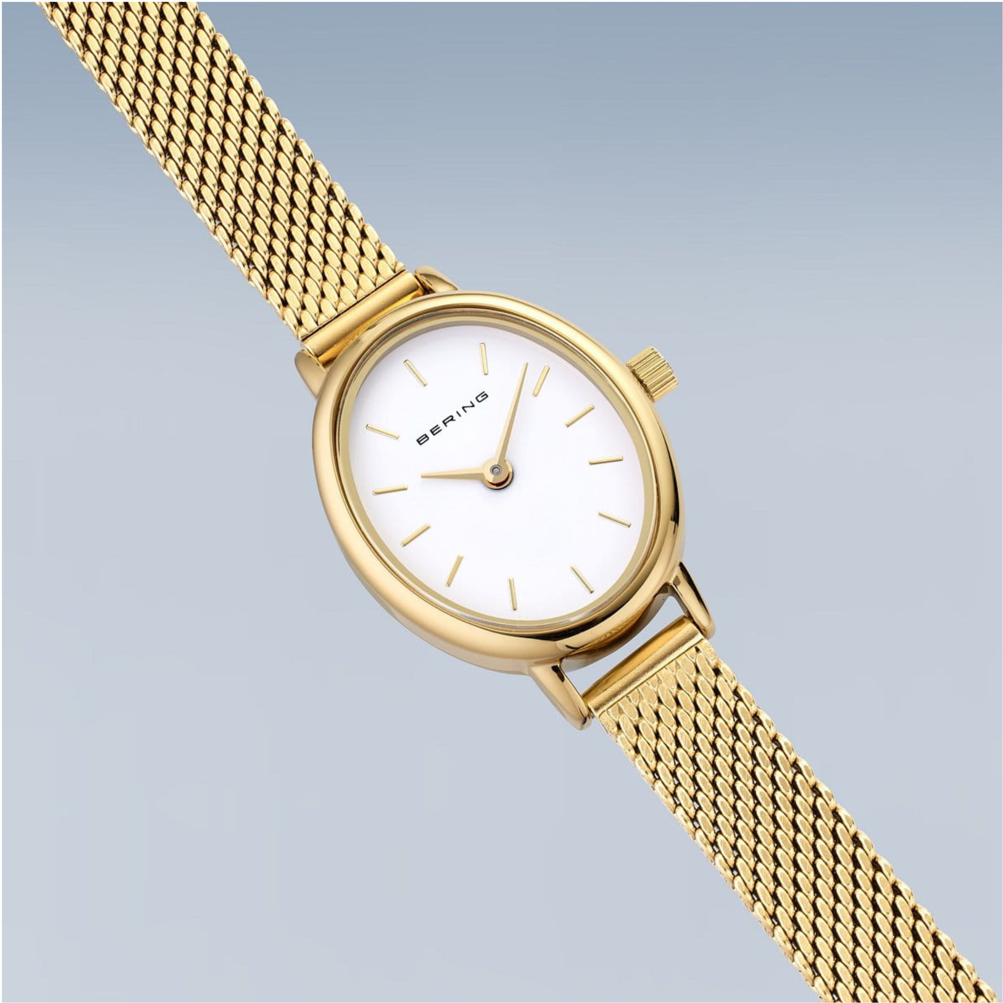 The Bering Watches Classic 11020-334 features a gold Milanese strap, white oval face, and minimal hour markers, shown diagonally on a light gradient background.