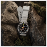The Luminox Watches MIL-SPEC XL.3359.SET by Luminox Watches, featuring a black dial and white strap, sits on a rocky surface with green moss. Bold numerals, luminous hands, and 300m water resistance ensure exceptional reliability anywhere.