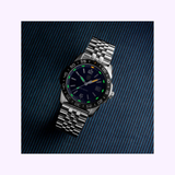 The Luminox Watches Pacific Diver Ripple XS.3123M.SET.1, featuring a stainless steel case, black dial, and metal link bracelet, is displayed at an angle against a dark, diagonally striped background.