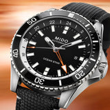 A close-up of the Mido Watches Ocean Star GMT M0266291705101, a Swiss automatic diver with black dial, silver markers, orange accents, and a black textured strap, set against a backdrop inspired by Miami sunsets.