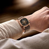 A person in a beige ribbed long-sleeve shirt wears the Mido Watches Multifort TV Big Date M0495263304100, featuring a brown and gold round face, analog hands, small digital display at the top, and a stylish silicone strap.