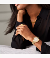 A woman in a black blouse rests her arm on a light-colored surface, highlighting a Michele Watches Serein Mid (Model: MWW21B000160, UPC: 99945535944) gold wristwatch by Michele Watches, complemented by a gold ring and gold necklace. Her face is partially out of frame.