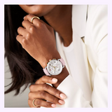 A person in a white blazer and gold ring showcases the MICHELE Watches SPORTY SPORT SAIL MWW01P000023, featuring a pale pink strap, silver face, and gold and silver accents from Michele Watches.