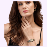A woman with long brown hair wears the Michele Watches Deco Madison Mini MWW06D000178 diamond watch in gold and silver, paired with gold earrings and a black spaghetti strap top, posing with one hand touching her neck against a plain light background.
