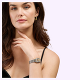 A woman with long brown hair, dressed in a sleeveless black top and gold hoop earrings, showcases the luxurious Michele Watches Deco Madison Mini MWW06D000178 diamond watch as she holds her hand near her chin against a light purple background.