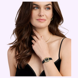 A woman with wavy brown hair is wearing a black spaghetti strap top, gold necklace, hoop earrings, and a Michele Watches Deco Madison Mini MWW06D000179 by Michele Watches on her wrist. She is looking off to the side with one hand near her neck.