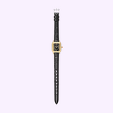 A Michele Watches Deco Madison Mini MWW06D000179 by Michele Watches, featuring a gold square case, white dial with black Roman numerals, and a sleek black leather strap, is displayed on a light purple background.