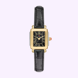 A Michele Watches Deco Madison Mini MWW06D000179 rectangular gold and silver wristwatch, featuring a black leather strap, black dial with Roman numerals, and a red accent on the crown, is displayed on a light purple background.