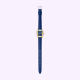 The Michele Watches Deco Madison Mini MWW06D000180, a gold rectangular timepiece with a blue face adorned with Roman numerals and set on a navy leather strap, is elegantly displayed against a light purple background.