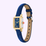 The Michele Watches Deco Madison Mini MWW06D000180 is a rectangular gold wristwatch featuring a striking blue face with Roman numerals and a navy leather strap, displayed on a light background.