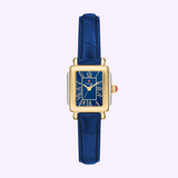 A Michele Watches Deco Madison Mini MWW06D000180 wristwatch featuring a rectangular gold case, a blue Roman numeral dial, and a navy leather strap, displayed on a light purple background.